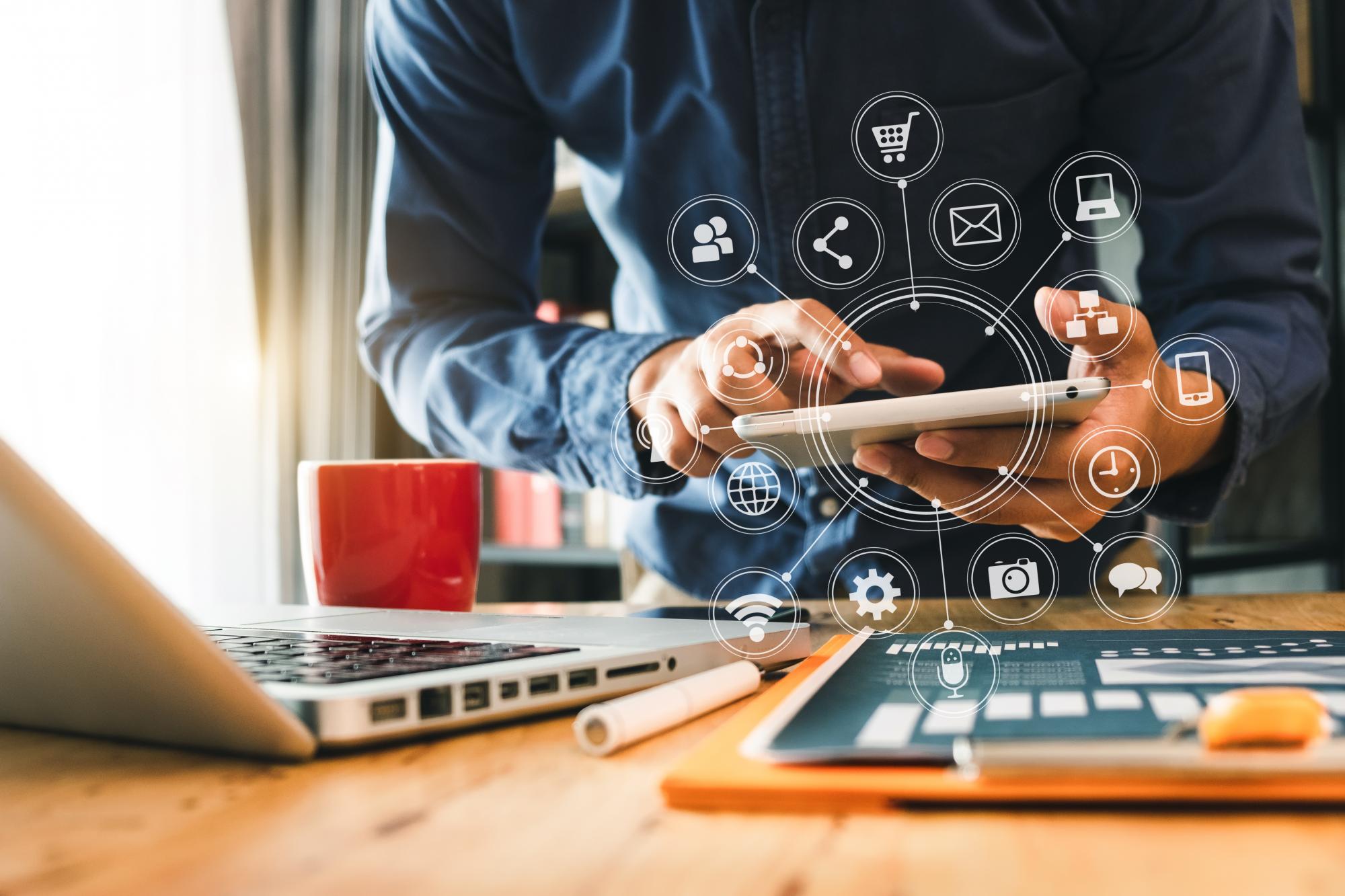 A busy employee works on a tablet armed with a laptop, a cup of coffee and a stack of papers. A series of icons indicating different services appears over the image. 